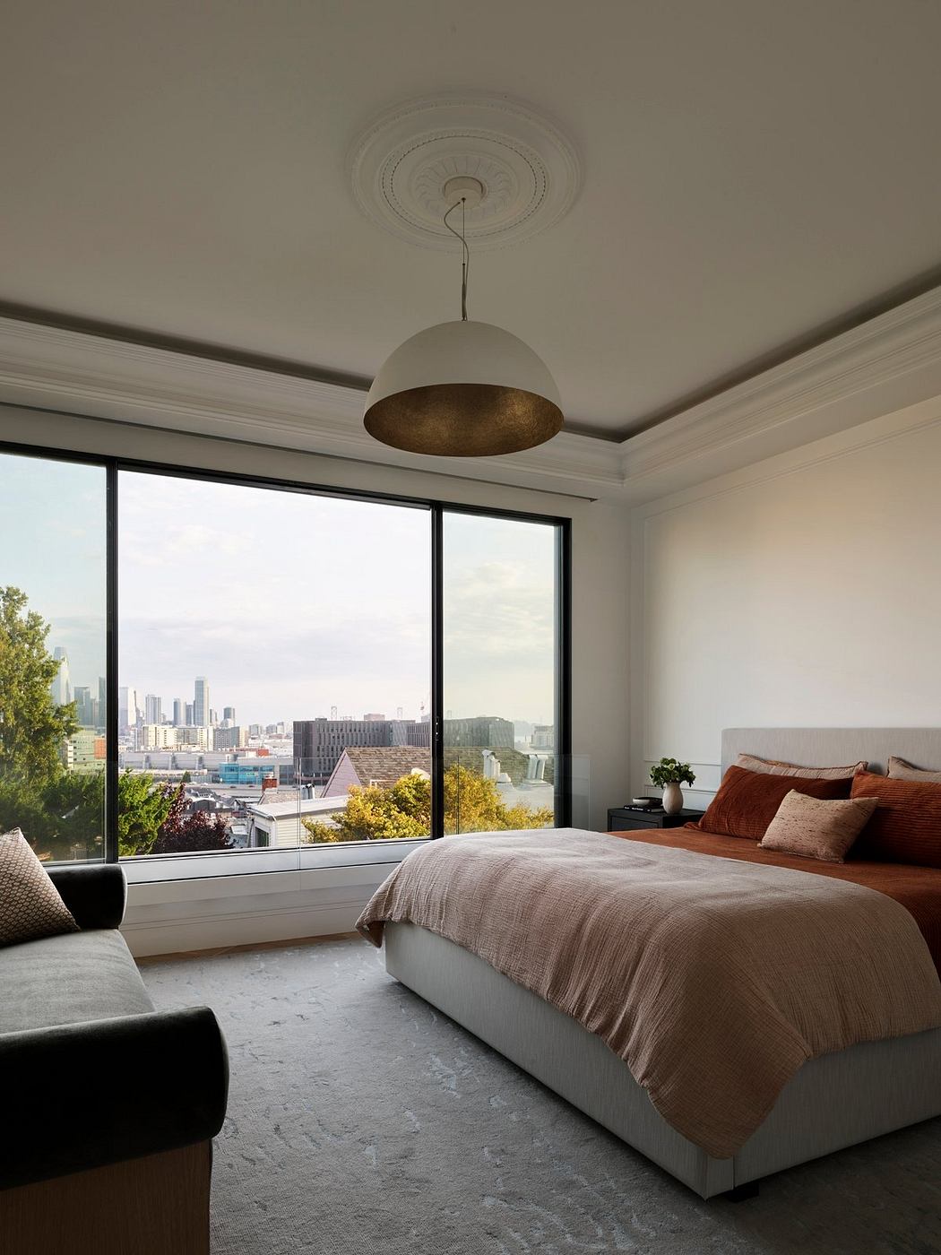 A cozy bedroom with a suspended lighting fixture, tall windows offering a cityscape view, and a plush rug.
