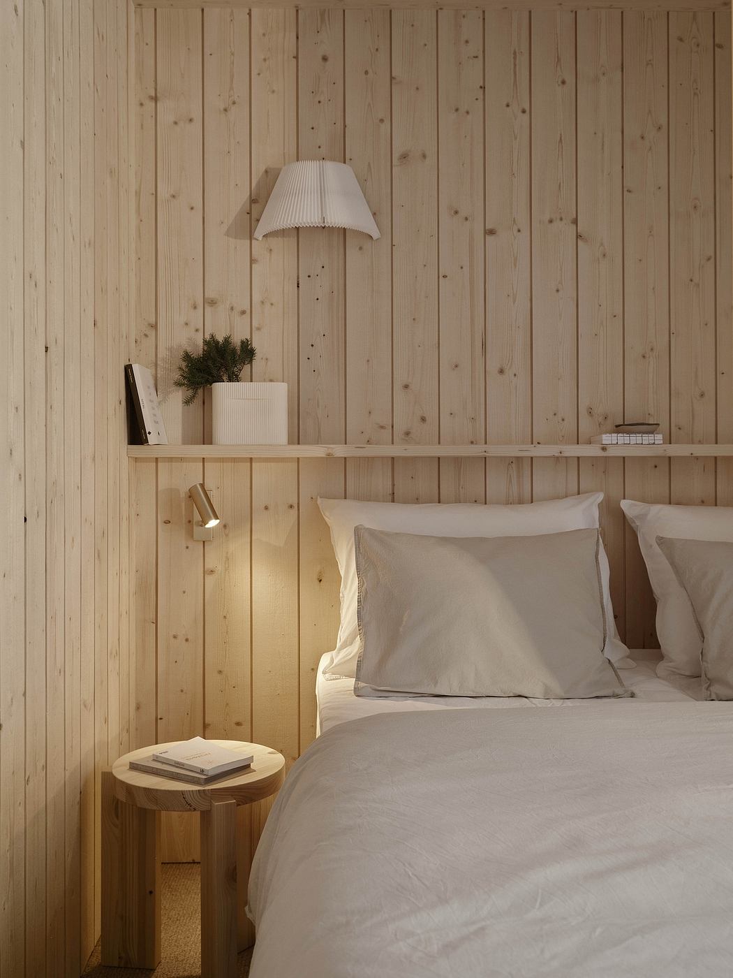 A cozy bedroom with a wooden wall, a lamp, and a bedside table displaying books and plants.