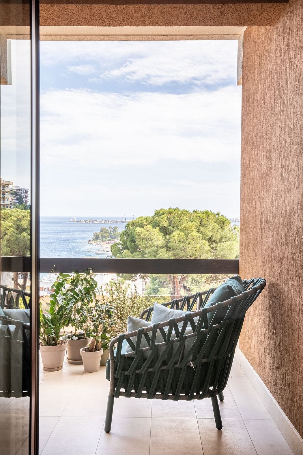 A cozy balcony with a woven chair, potted plants, and a scenic view of the cityscape beyond the glass door.