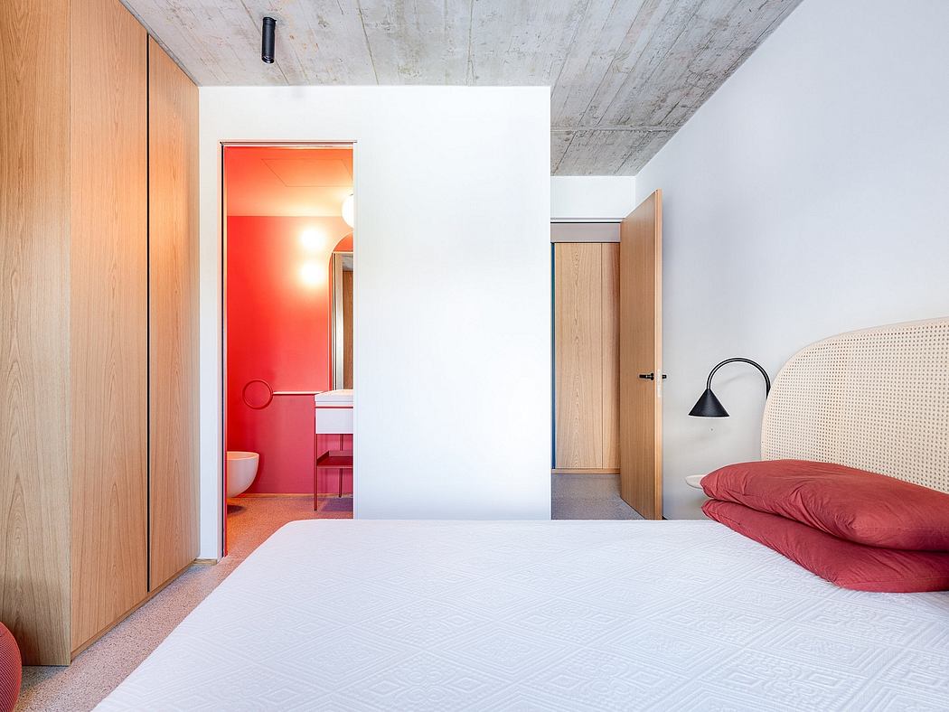 A minimalist bedroom with a bold red bathroom visible through an open doorway, concrete ceiling, and wooden accents.