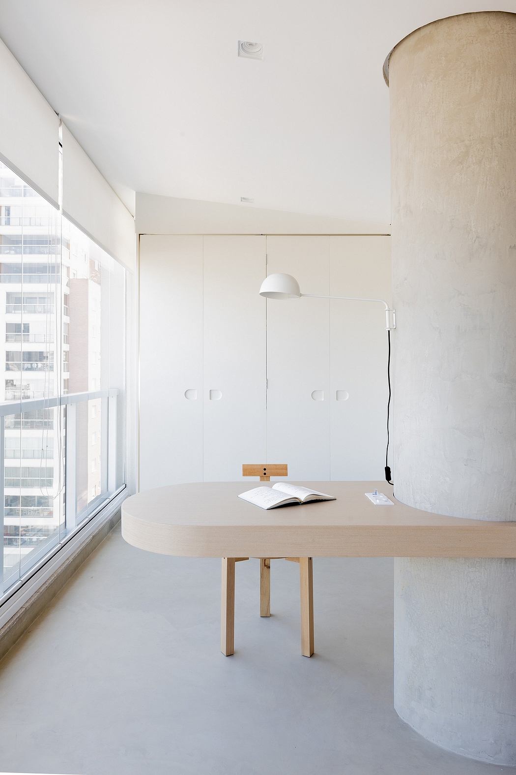 Minimalist office space with a round wooden table, neutral-toned walls, and a large window.