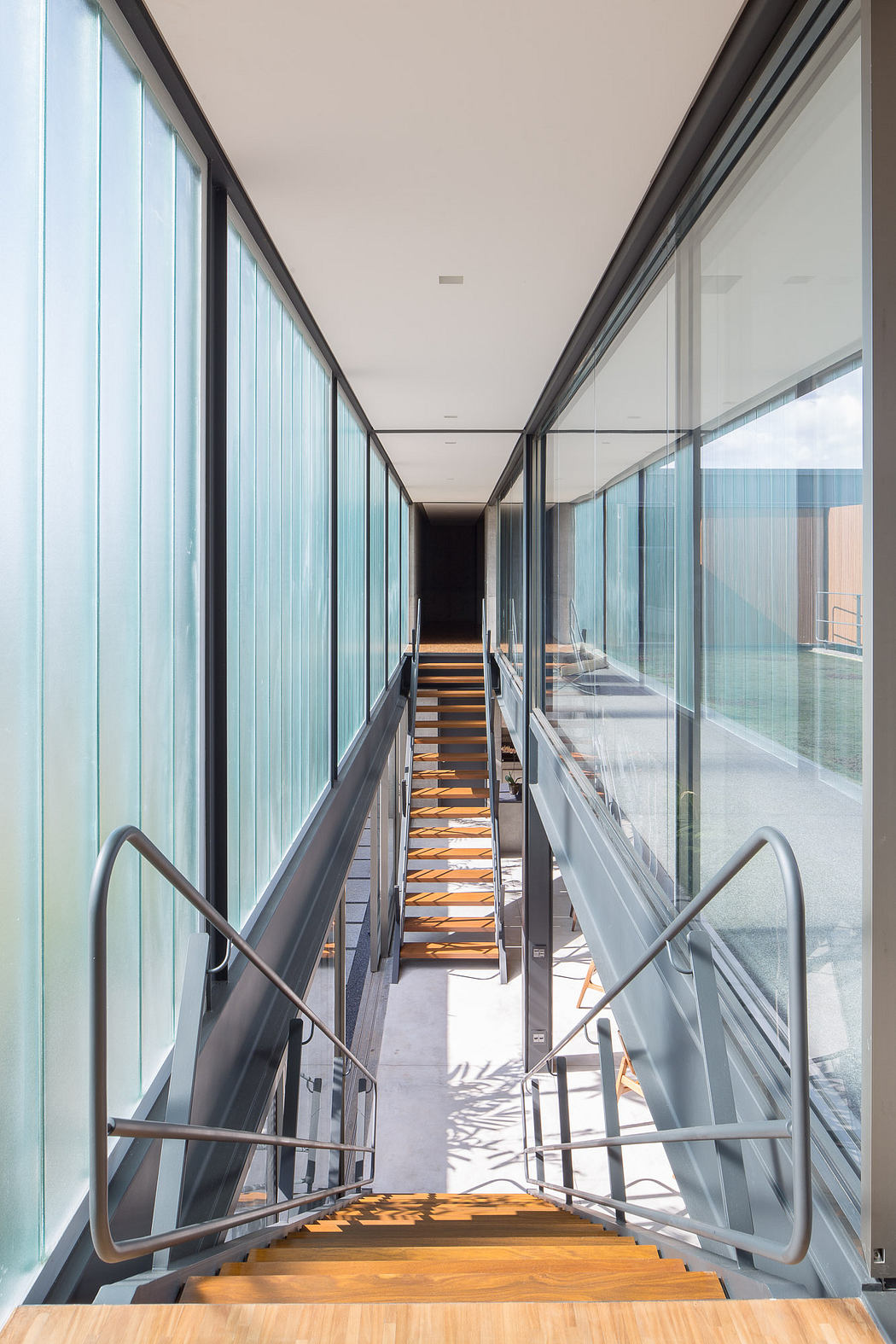 A modern interior hallway with glass walls, wooden stairs, and metal railings.