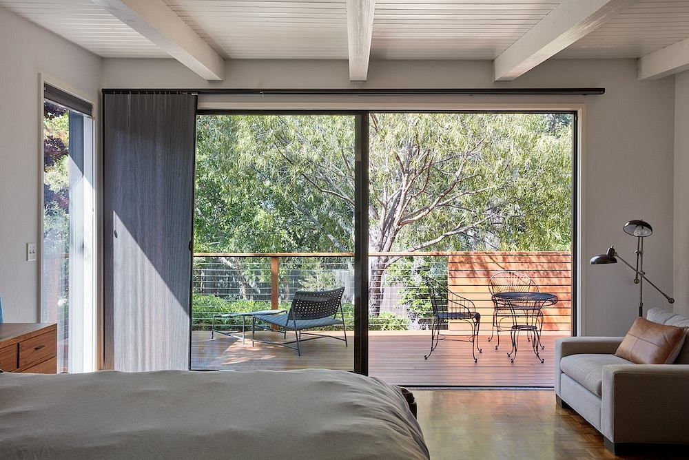 Cozy bedroom with large sliding glass doors overlooking lush greenery and outdoor seating.