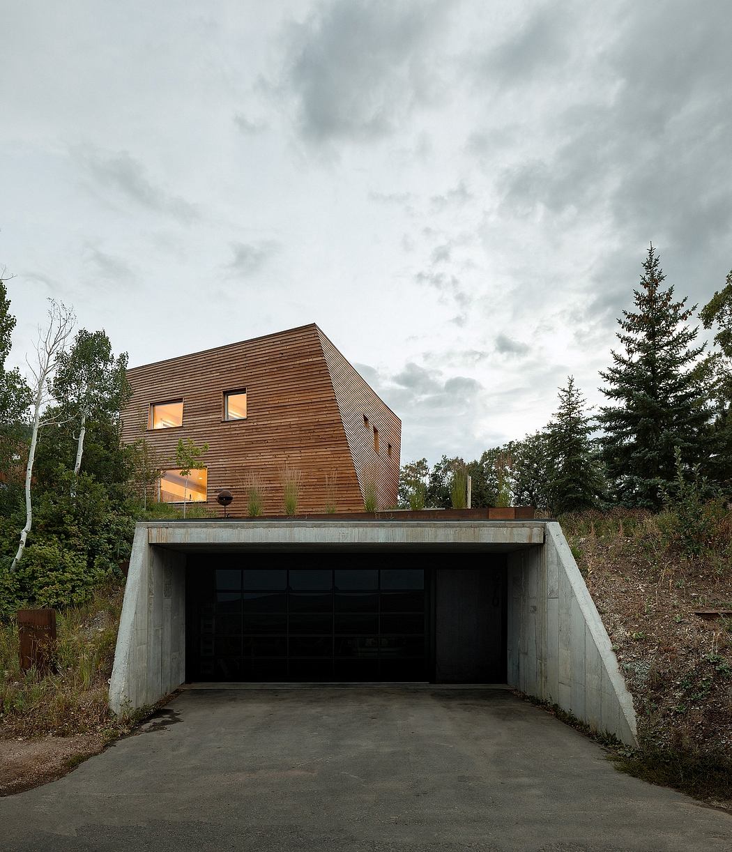 A modern, wooden-clad house with a concrete garage entrance and surrounding forest.