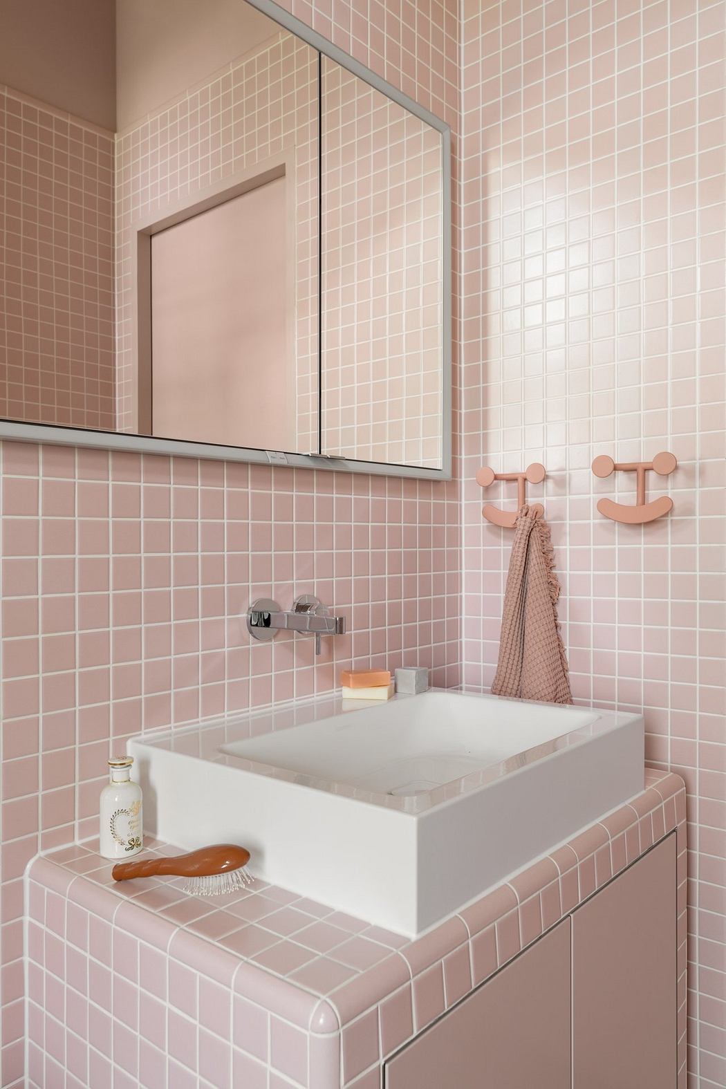 A pink-tiled bathroom with a rectangular white sink, wall-mounted mirror, and decorative hooks.