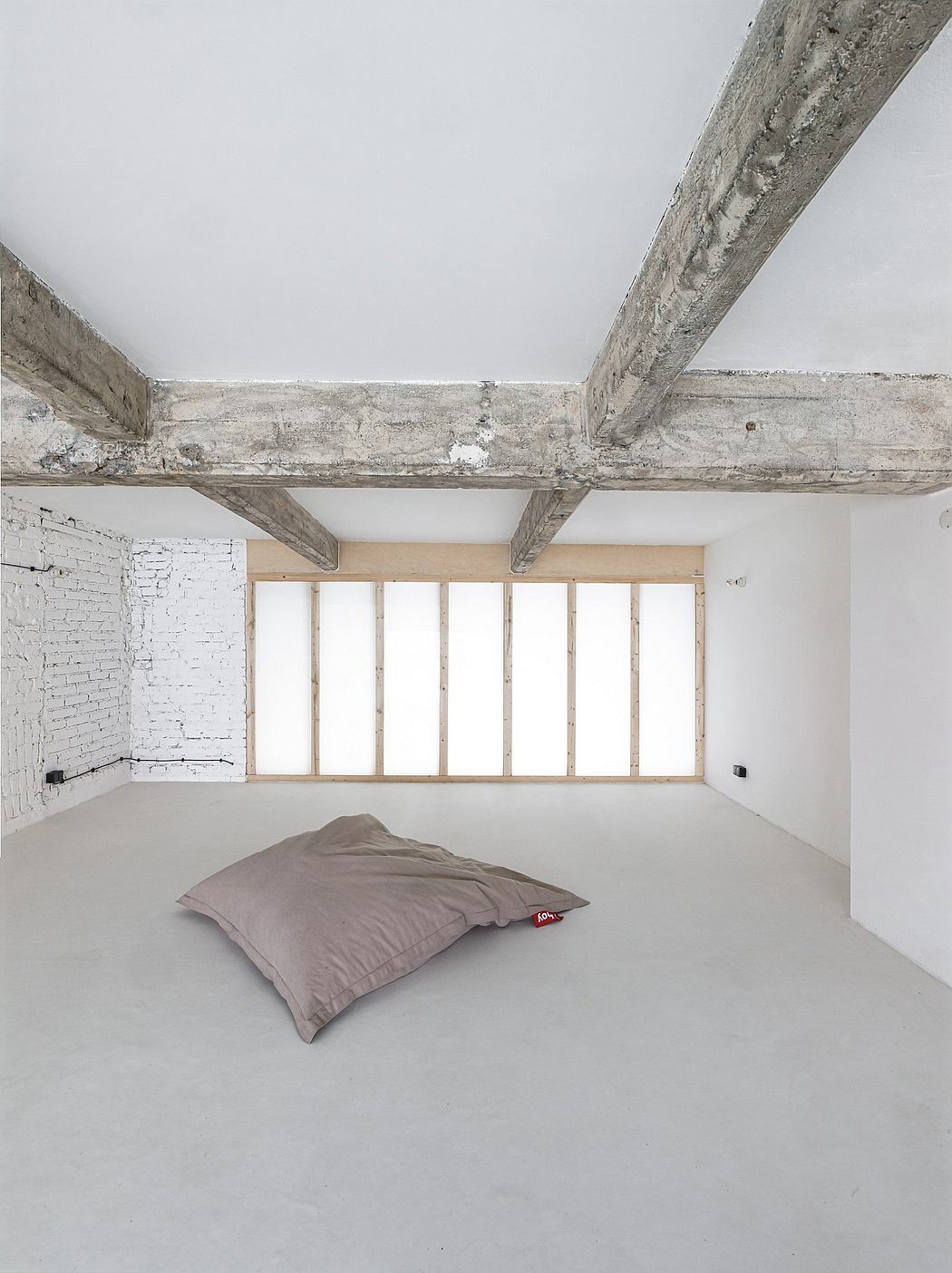 A minimalist loft-like space with a concrete ceiling, wooden beams, and an oversized floor cushion.