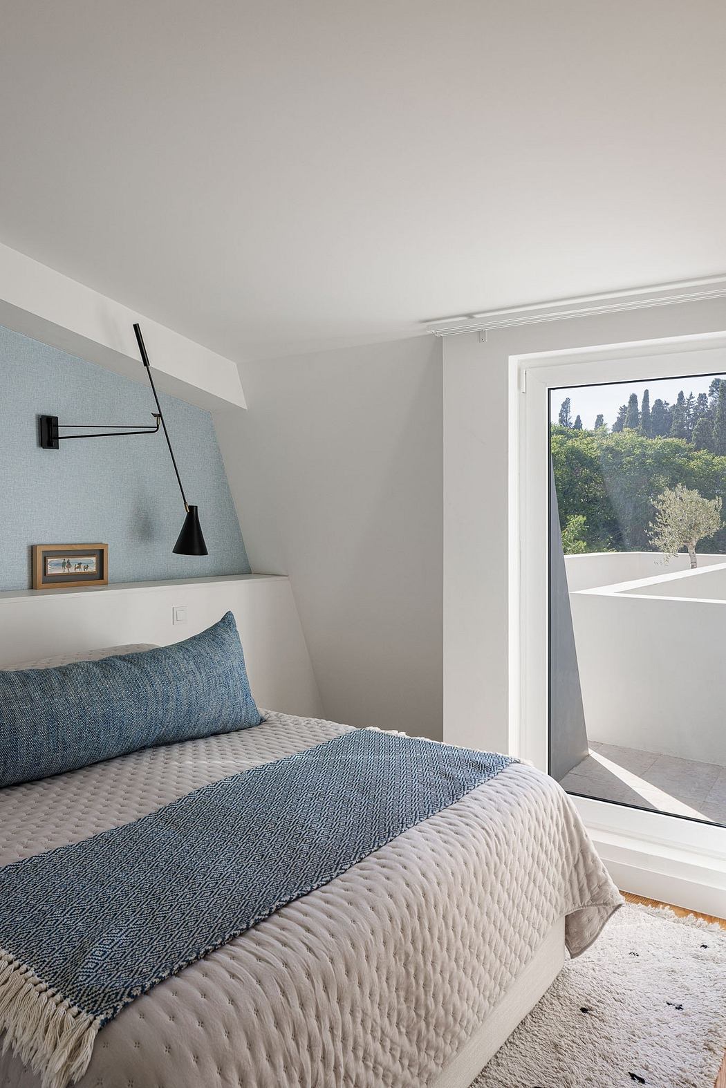 A cozy, minimalist bedroom with a large window, textured bedding, and a modern wall lamp.