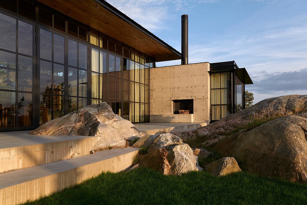 A modern, wood-clad building with glass walls, stone steps, and a tall chimney stands against a rugged landscape.