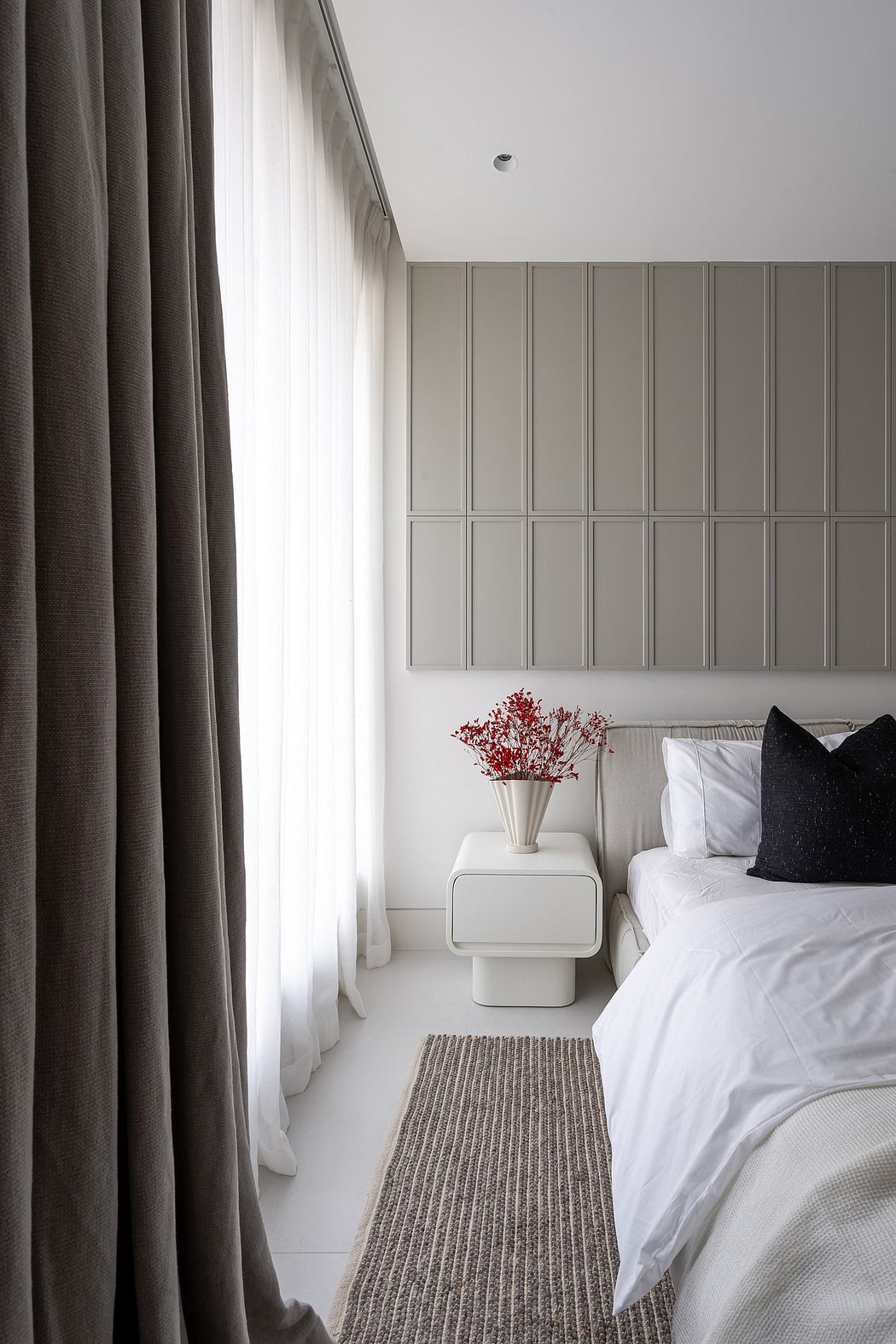 A cozy bedroom with gray curtains, a white bed, and a vibrant floral arrangement on a side table.