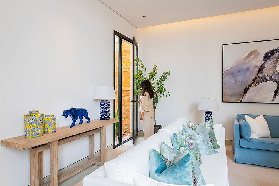 Modern, minimalist living room with wooden console, blue accents, and large window overlooking plants.
