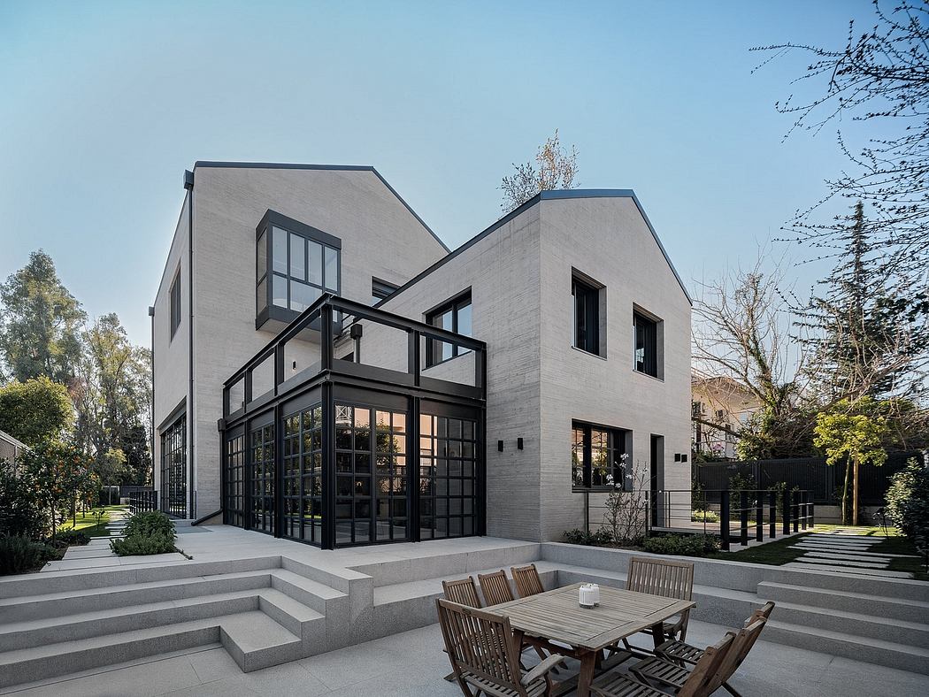 A modern, angular house with large windows, a wooden dining set, and lush greenery surrounding it.