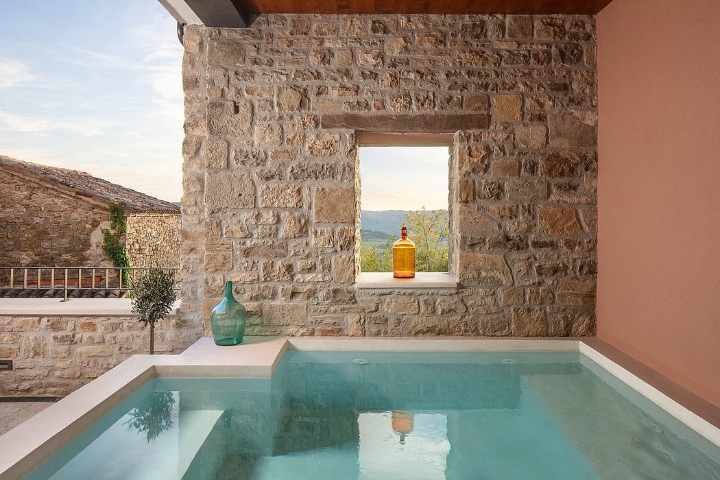 A stone-walled room with a window overlooking a swimming pool and scenic landscape.