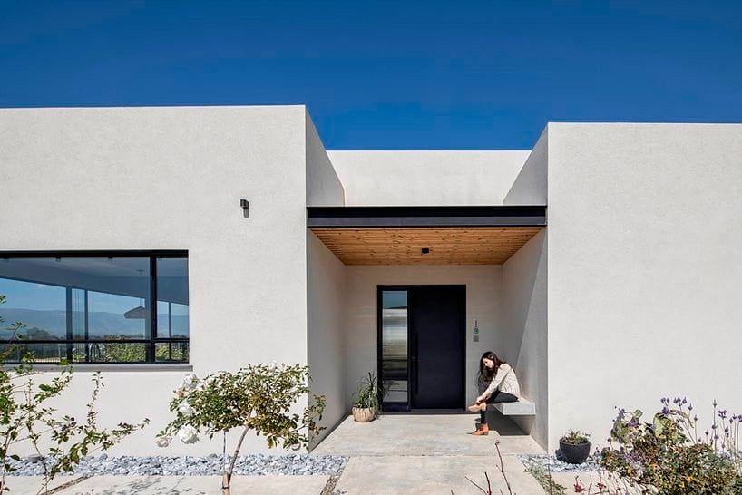 The image depicts a modern, minimalist home exterior with clean lines, wood-paneled entrance, and lush landscaping.
