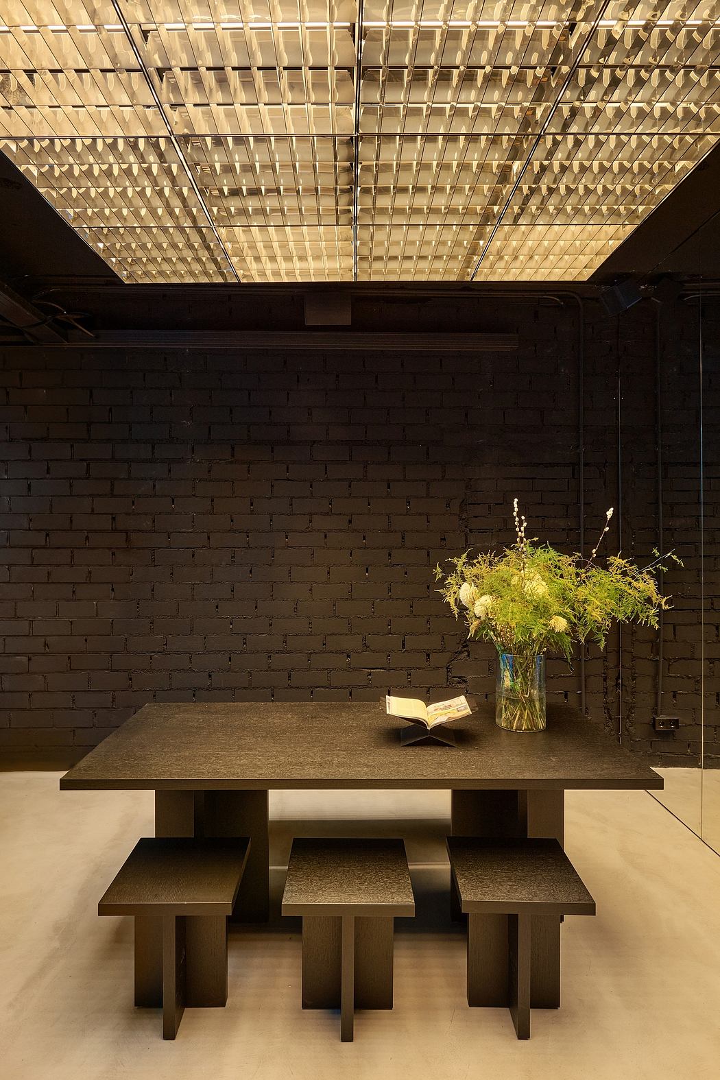 Minimalist dining area with geometric wooden ceiling panel, dark brick walls, and a simple wood table with a vase of flowers.