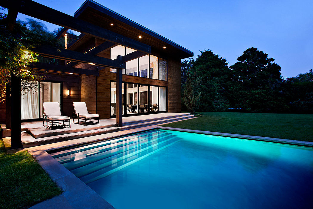 A modern, wooden house with a large pool, surrounded by lush greenery at night.