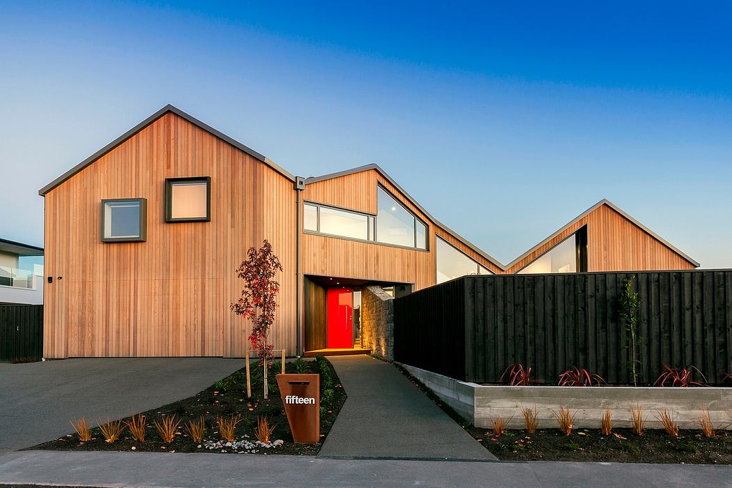 A modern wooden building with a red front door, surrounded by landscaping and a black fence.