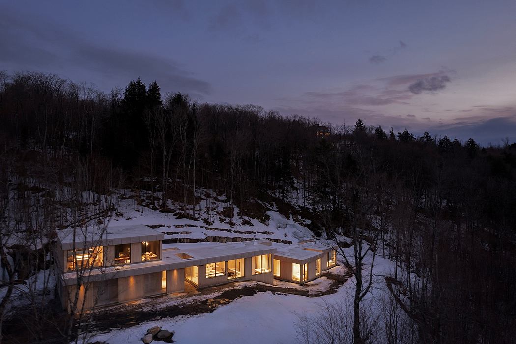 A modern, glass-walled cabin nestled in a snowy forest, illuminated at dusk.