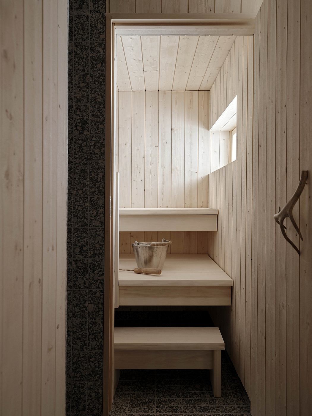 Rustic wooden sauna interior with bench and metal bucket on tiled floor.