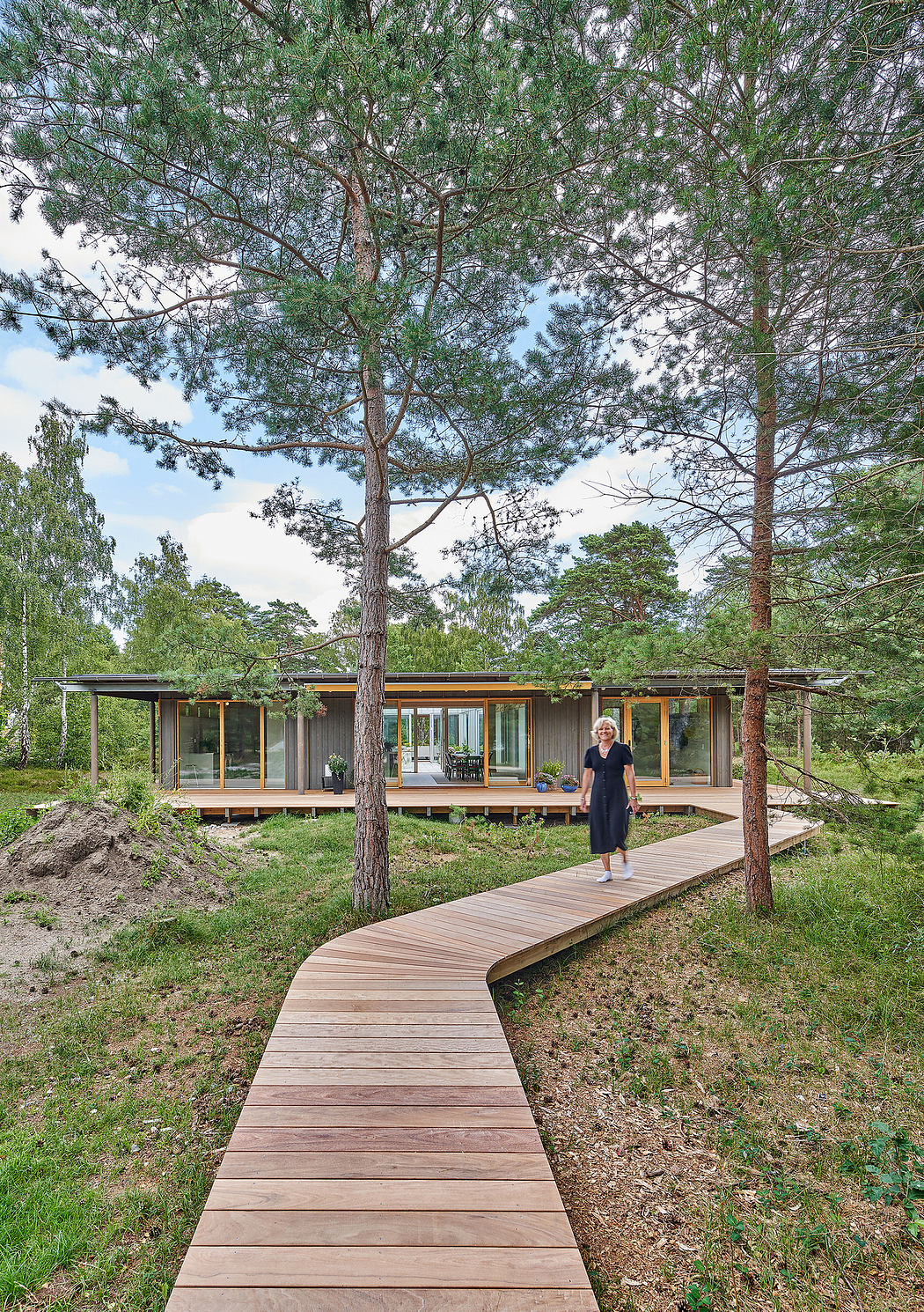 A single-story modern cabin surrounded by a lush pine forest, with a long wooden walkway leading to the entrance.