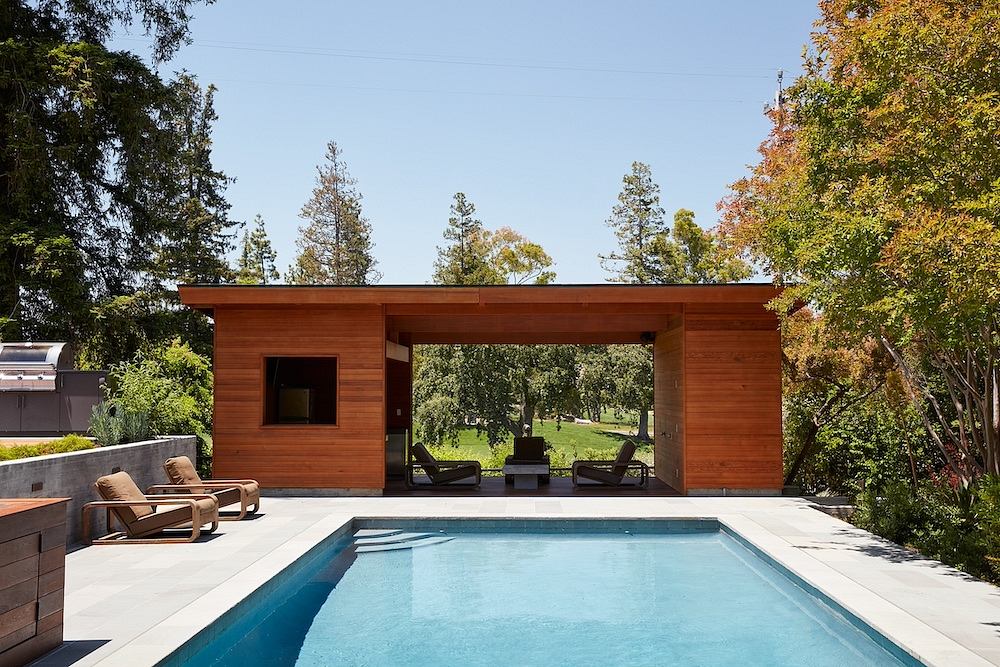 Modern wooden poolhouse with seating area, overlooking lush greenery.