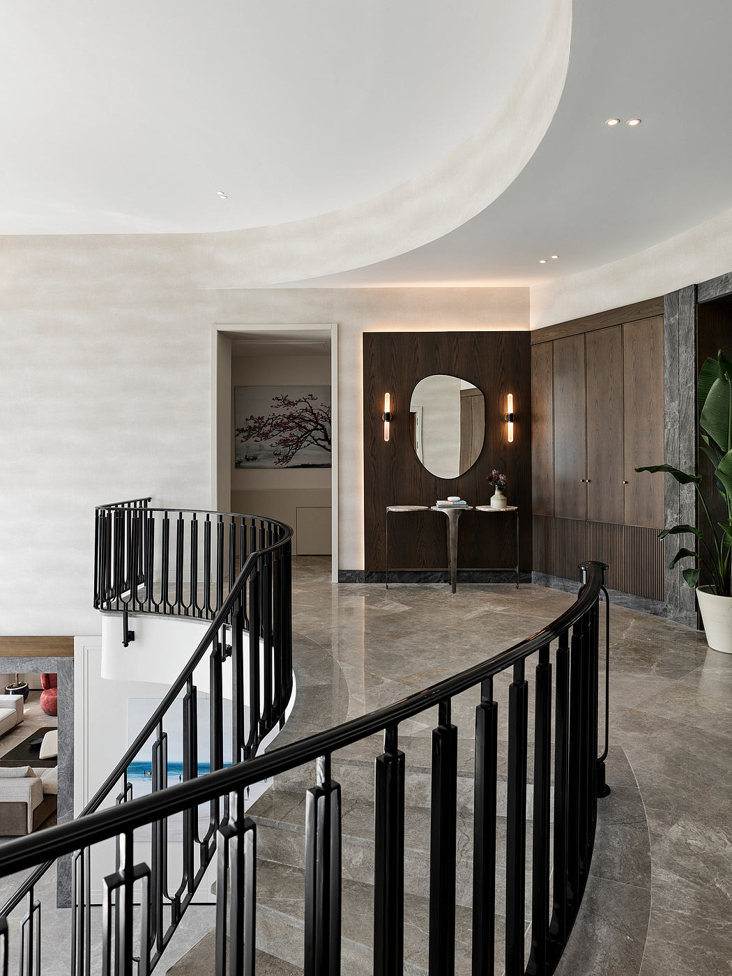 Elegant foyer with curved staircase, wood paneling, and oval mirror accented by wall sconces.