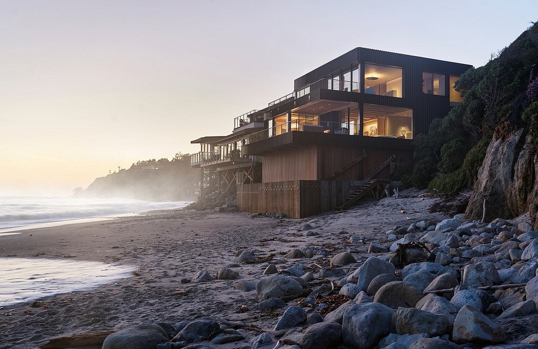 A modern, multi-level beachfront home with expansive decks, large windows, and a rocky shore.