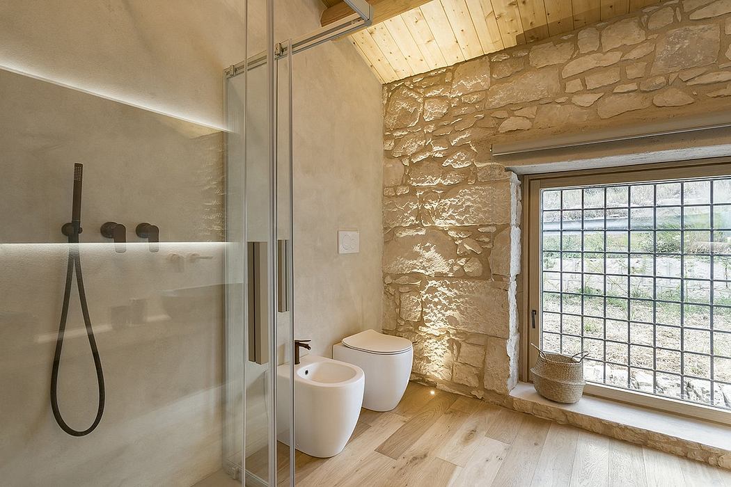 A rustic bathroom with stone walls, wooden ceiling, and large window overlooking the outdoors.