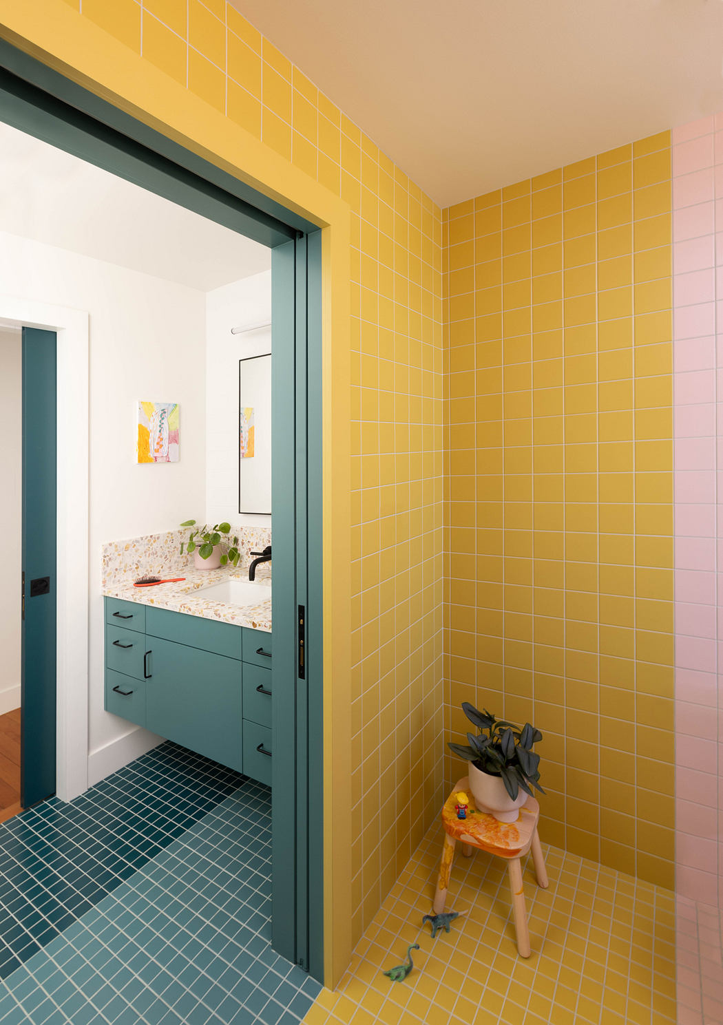 Vibrant yellow tiles cover the walls and floor, contrasting with the teal vanity and decor.