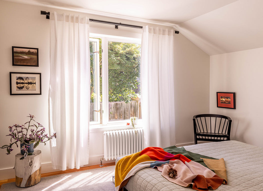 A cozy bedroom with large white-curtained window, radiator, and colorful textiles.