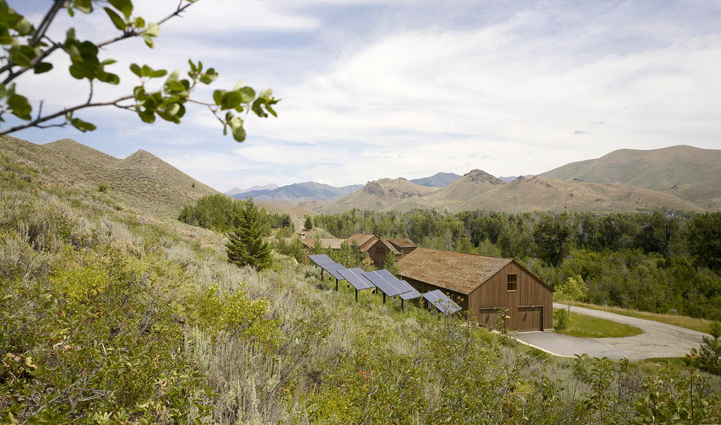 Rustic cabin amidst rugged mountains, solar panels on rooftop, surrounded by lush vegetation.