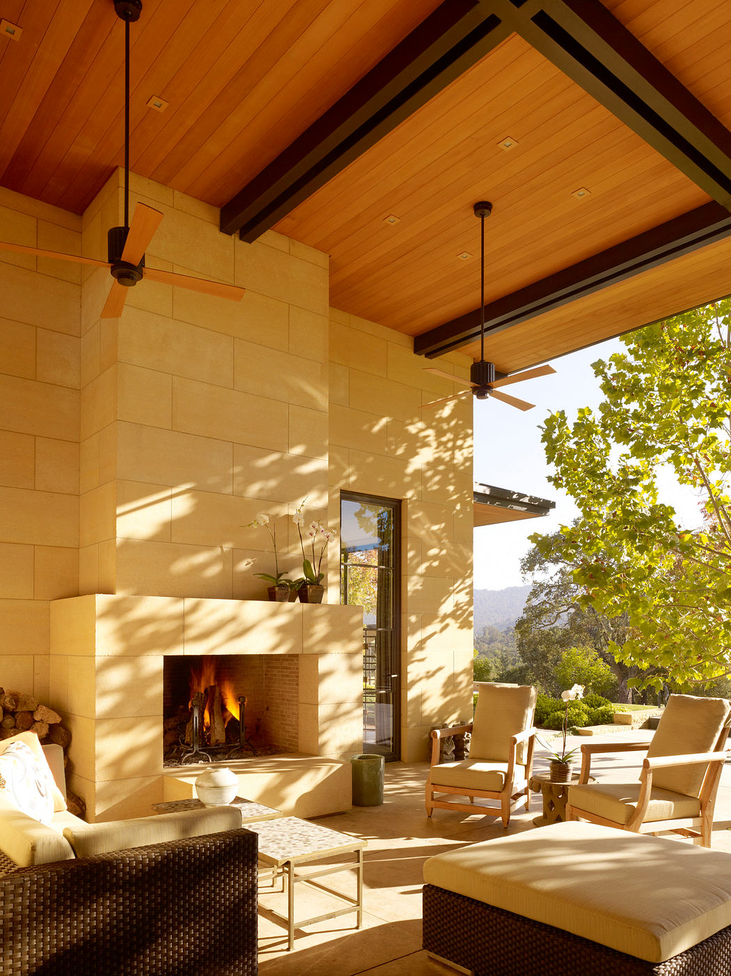 Warm, cozy outdoor lounge with fireplace, wood-paneled ceiling, and lush greenery.