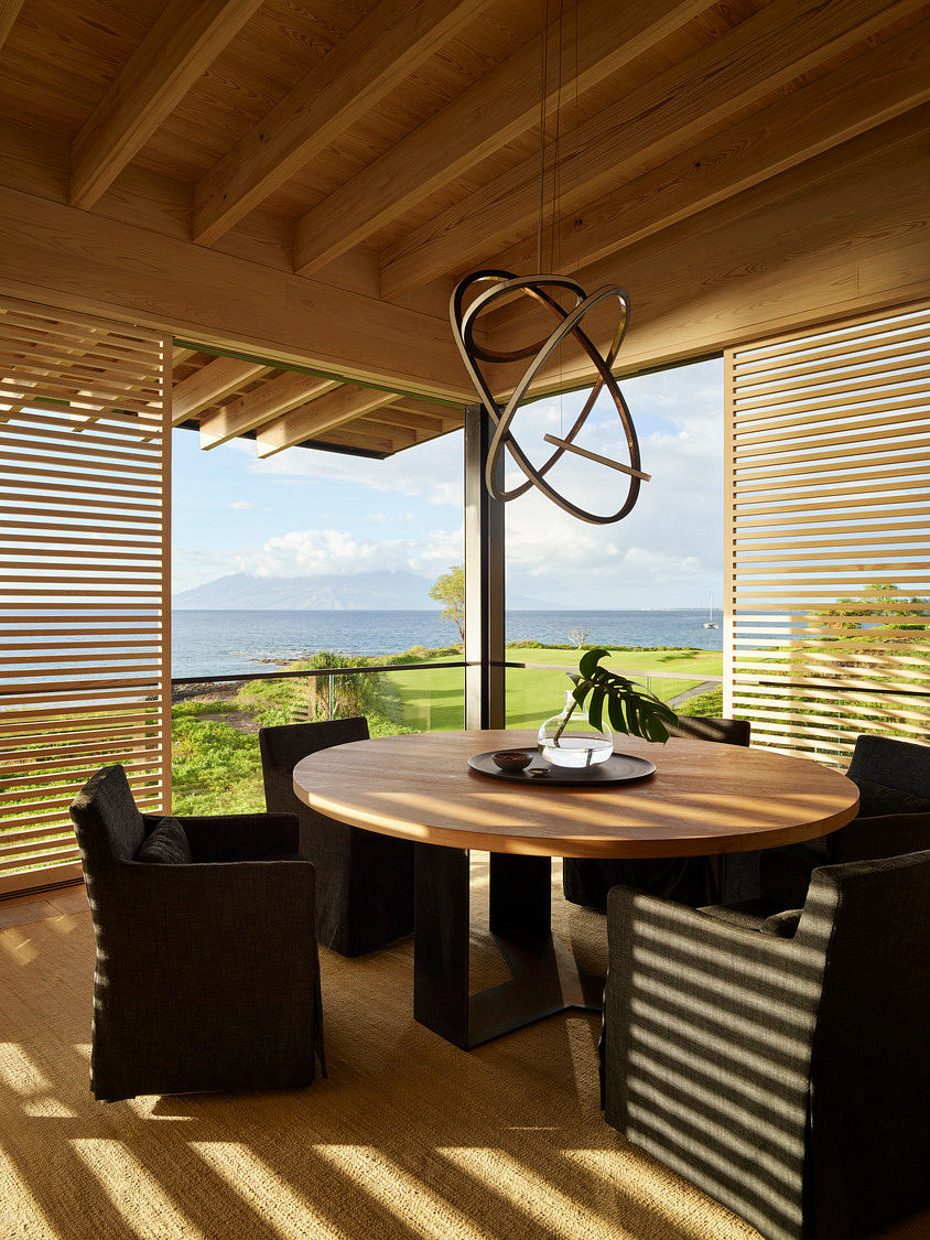 A modern, open-concept dining area with a round wooden table, black chairs, and a unique light fixture overlooking the ocean.