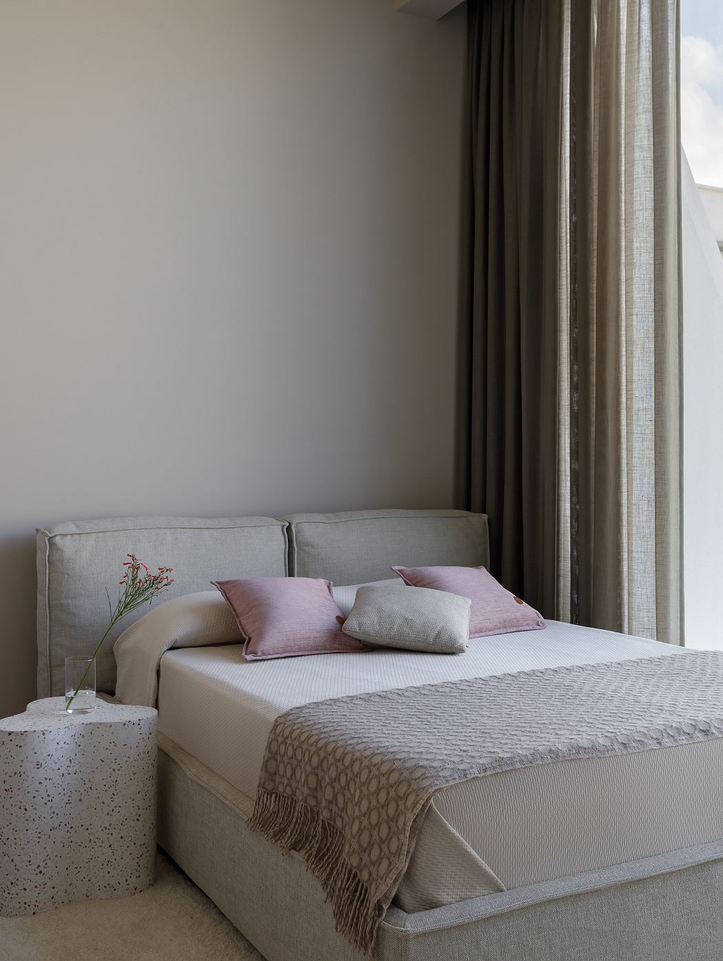 A cozy bedroom with a plush gray bed, soft throw, and subtle floral accents. Elegant curtains frame a window.