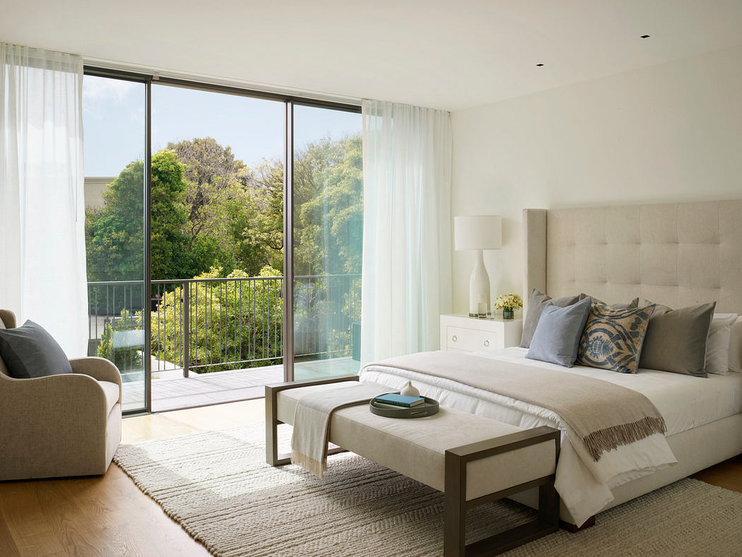Spacious, light-filled bedroom with large windows, plush furnishings, and lush greenery visible outside.