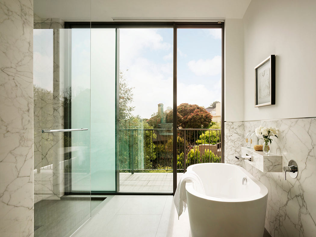 Spacious bathroom with marble walls, freestanding tub, and expansive glass windows overlooking garden.