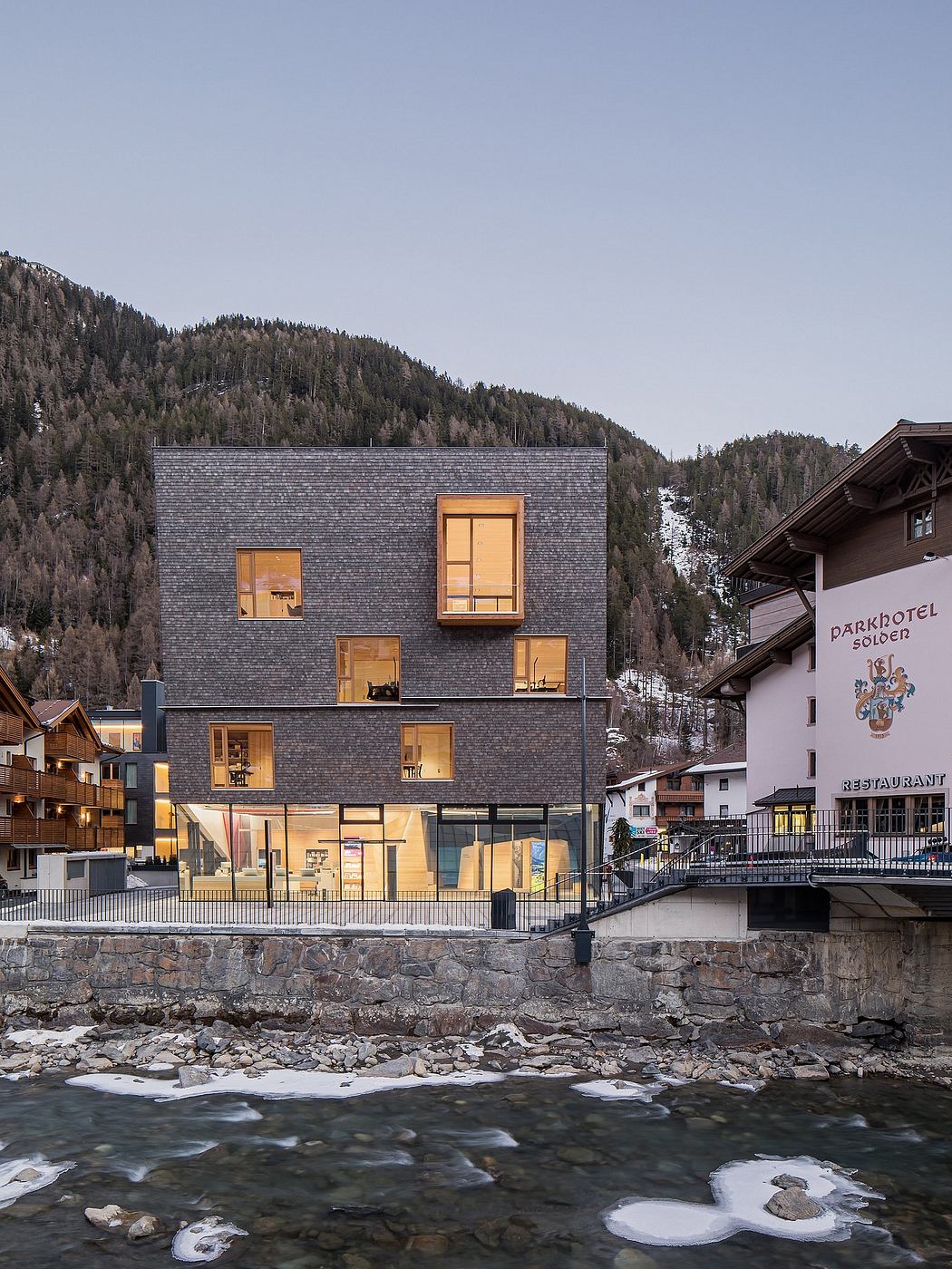 A modern, four-story building with a slate-gray exterior and large windows overlooking a snowy mountain landscape.