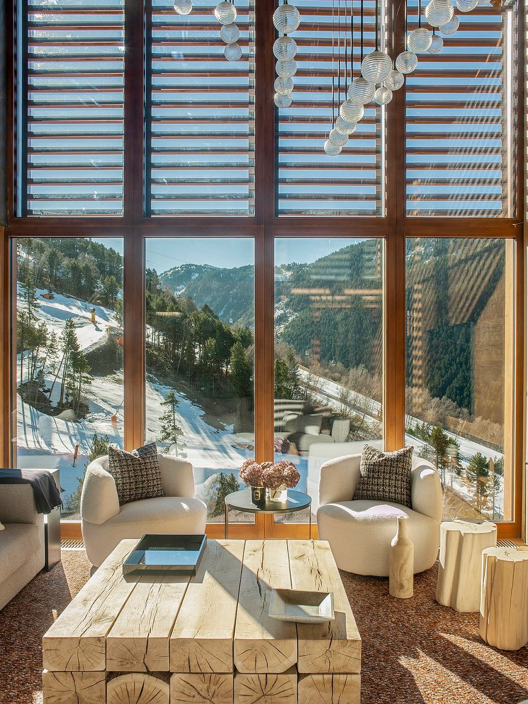 Cozy living area with wooden furniture, glass walls overlooking snowy mountains.
