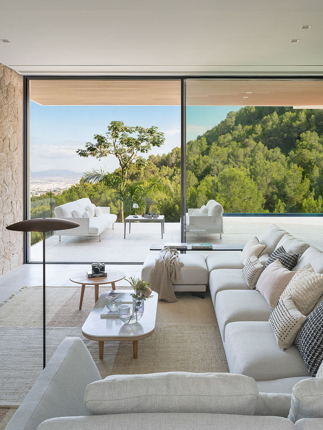 Expansive glass walls frame a lush, forested landscape beyond a serene living space.