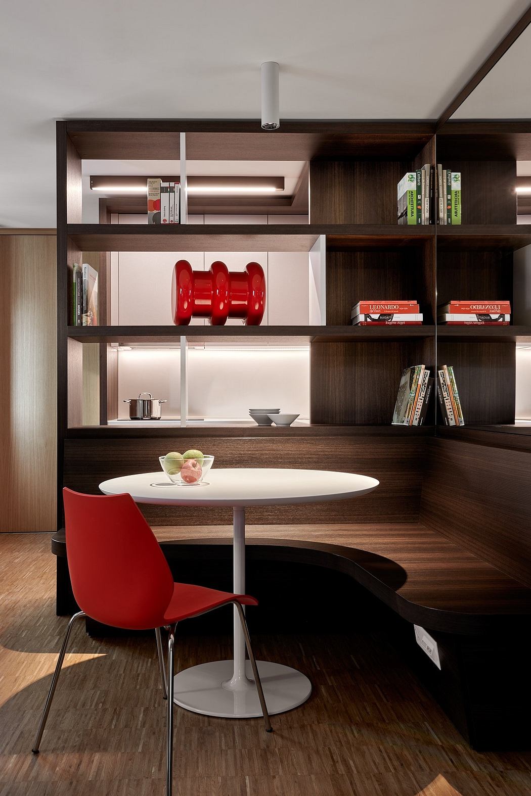 Minimalist dining nook with sleek wood shelving, white table, and vibrant red chairs.