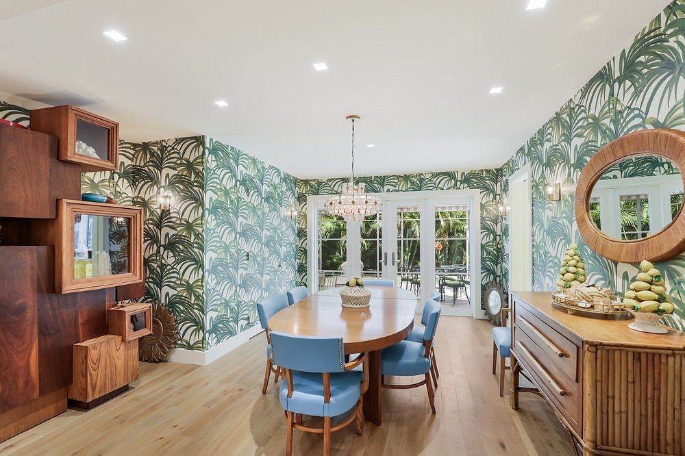 Vibrant tropical wallpaper, round mirror, wooden furniture, and large chandelier create a stylish dining space.