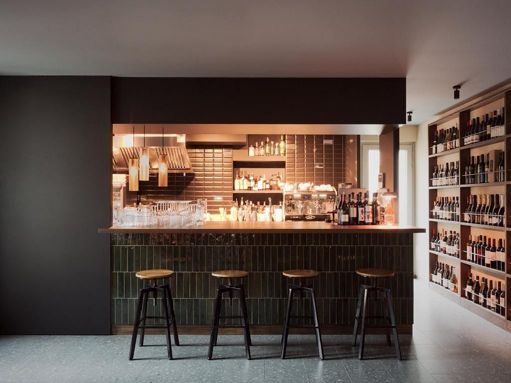 A stylish bar interior with a sleek black tiled counter, shelves displaying bottles, and wooden stools.