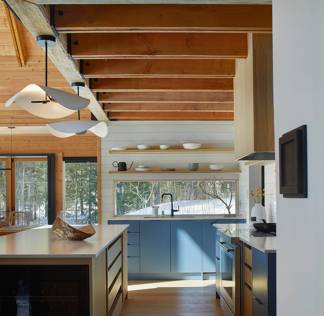 Warm, modern kitchen with wooden beams, shelving, and blue cabinets overlooking snowy trees.
