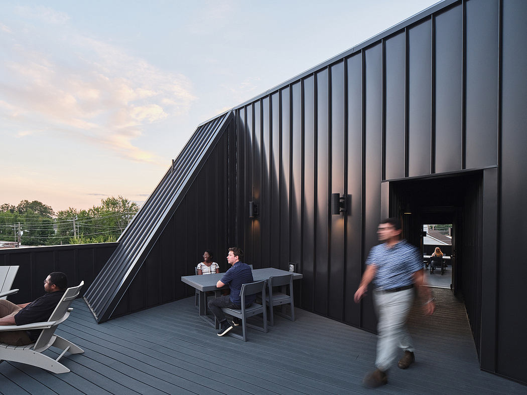 Modern black building with an angled facade, an outdoor deck, and people seated at a table.