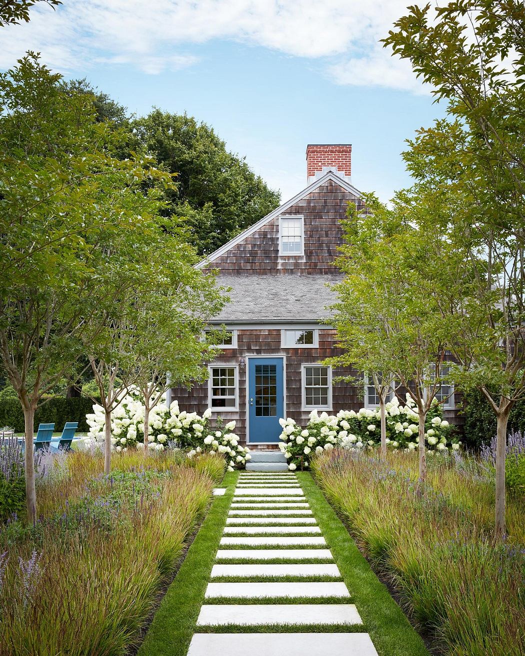 A charming coastal cottage with shingle siding, a blue front door, and a lush, manicured garden pathway.