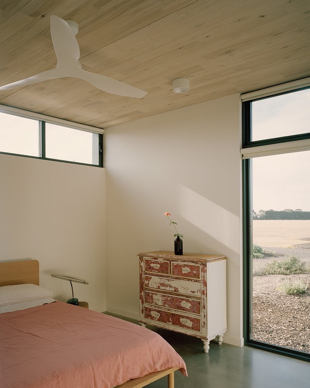 A minimalist bedroom with a wooden ceiling, a distressed dresser, and a panoramic window overlooking nature.