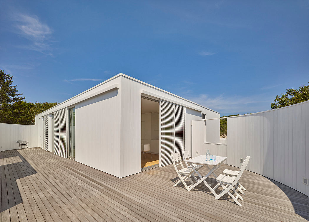 A modern, minimalist cabin with a large deck and outdoor seating area, surrounded by lush greenery.