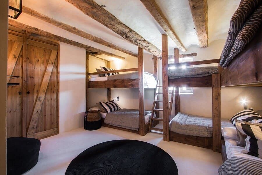 Rustic, cozy bunk room with exposed wooden beams, ladders, and textured walls.