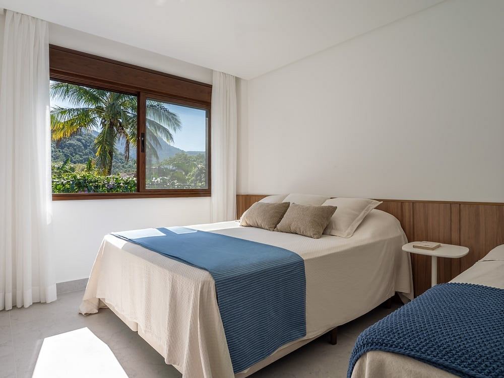 Serene bedroom with large window showcasing lush tropical landscape. Cozy furnishings, wood paneling.