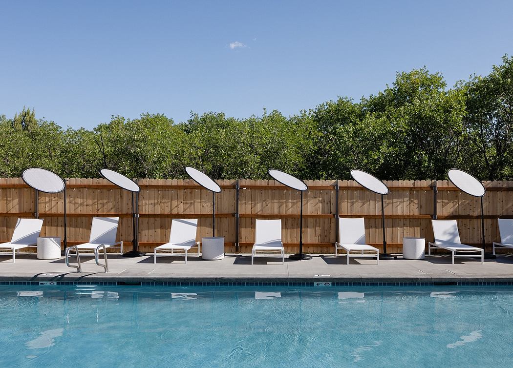 Sleek, modern pool area with row of white lounge chairs and shade umbrellas against wooded backdrop.