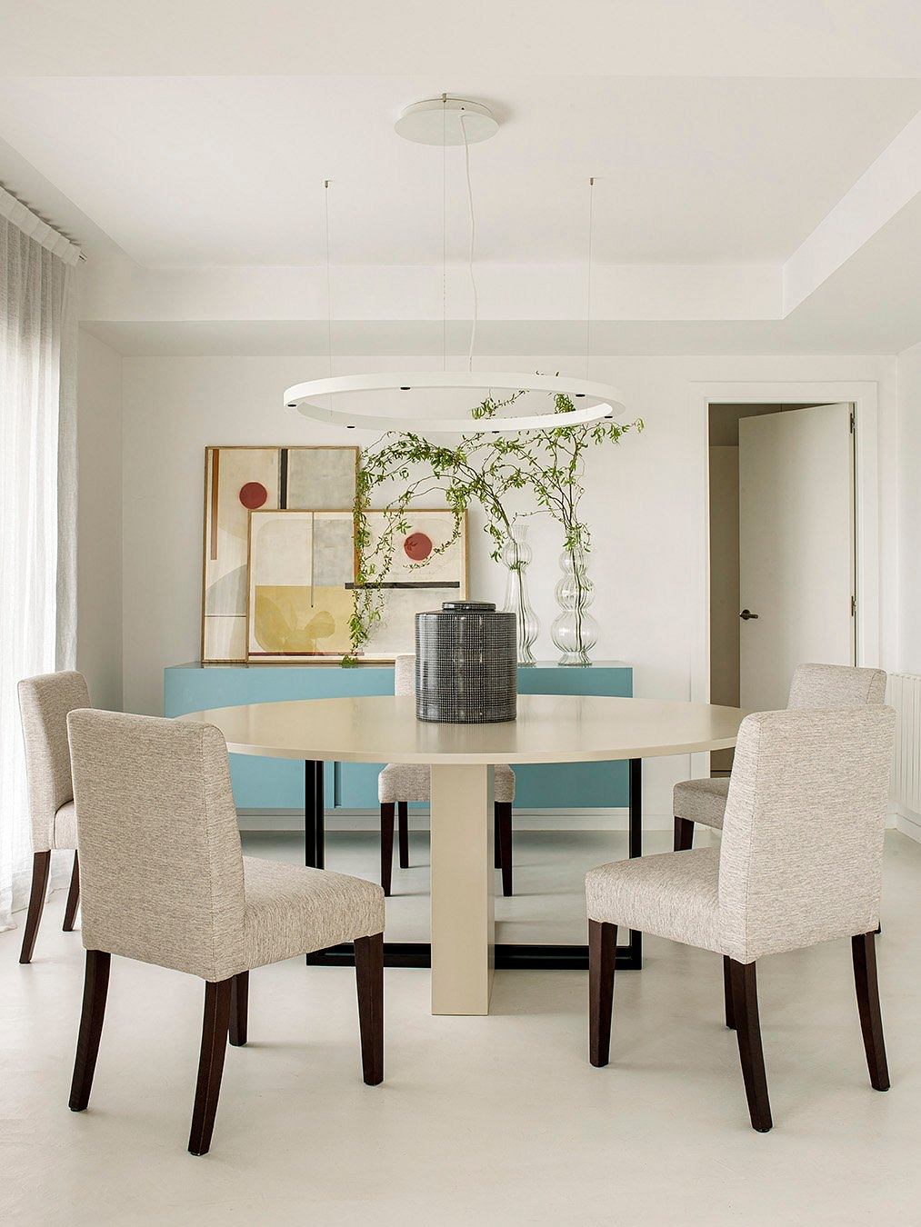 A modern, minimalist dining room with a round table, upholstered chairs, and a hanging light fixture.